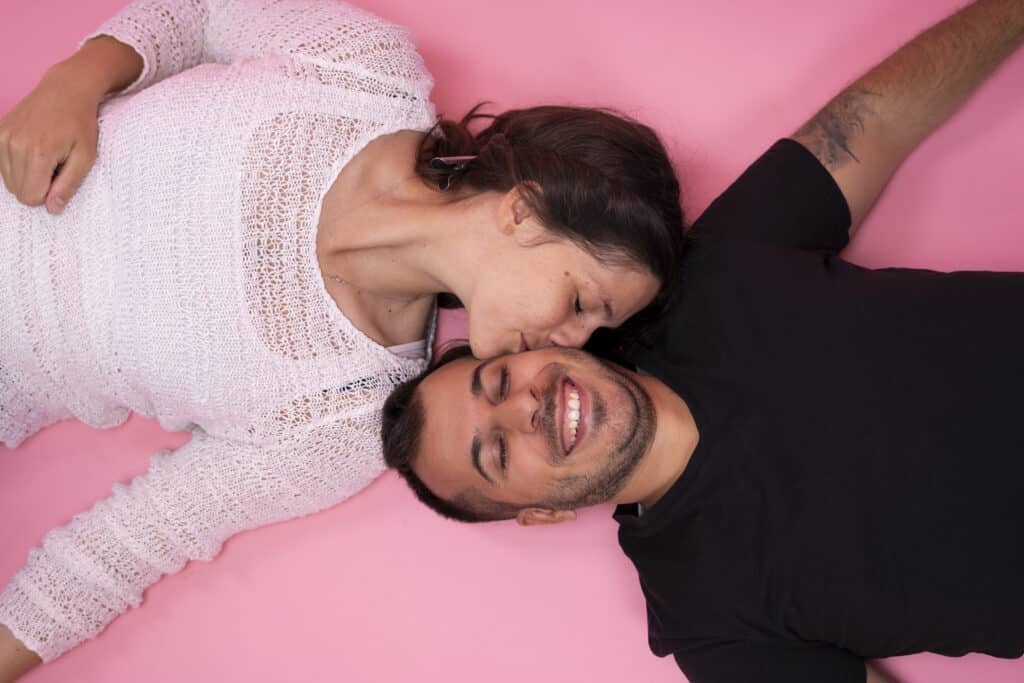 Glückliches Paar liegt eng beieinander und lacht vor einem pinken Hintergrund.