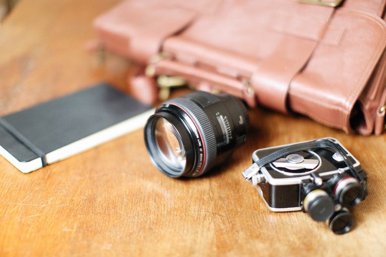 A vintage camera and lens rest on a table next to a leather bag and notebook, perfect for photography enthusiasts.